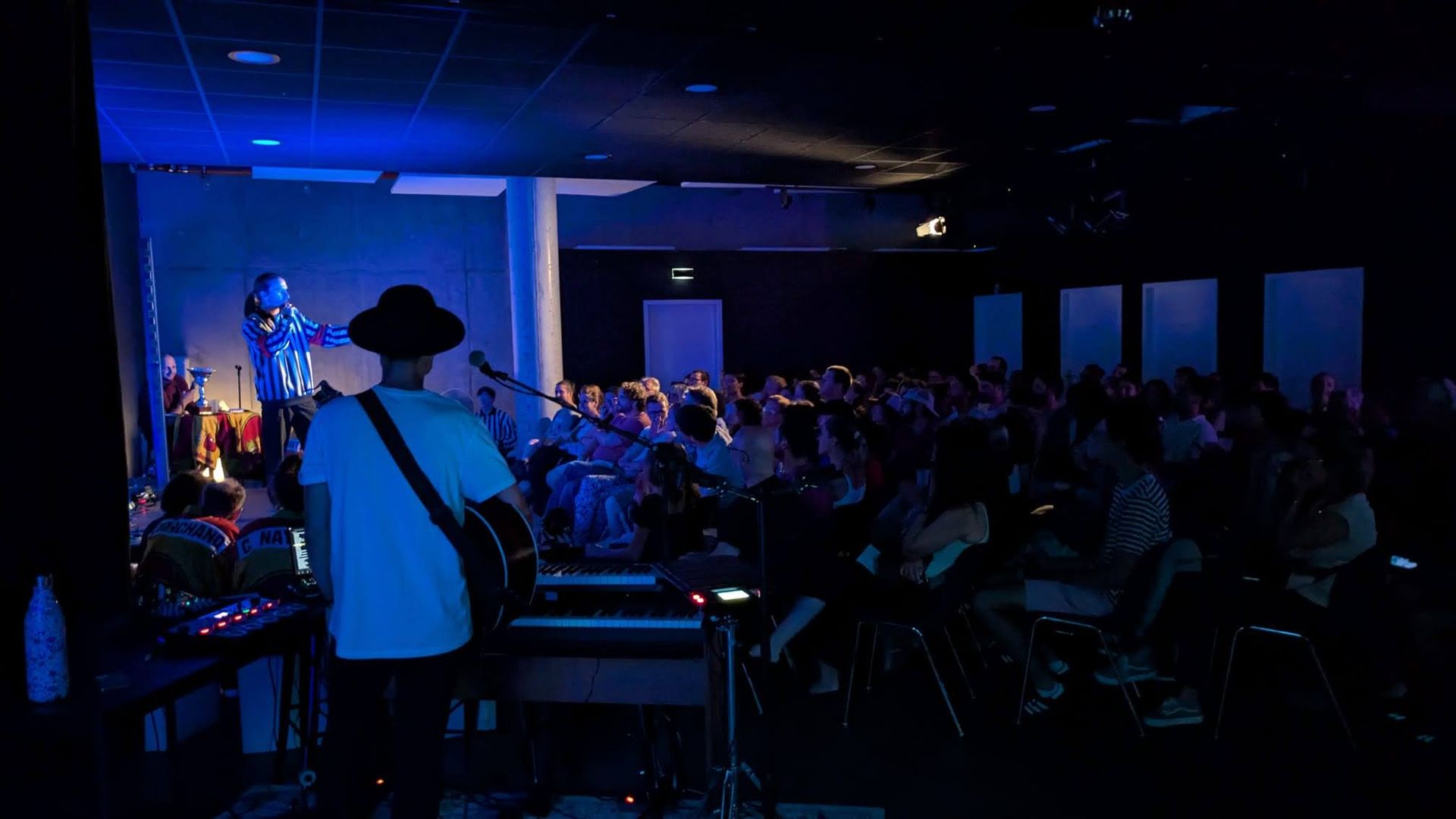 Un spectacle à L'Estrade : un humoriste sur scène sous les lumières bleues, un musicien à la guitare et la salle comble